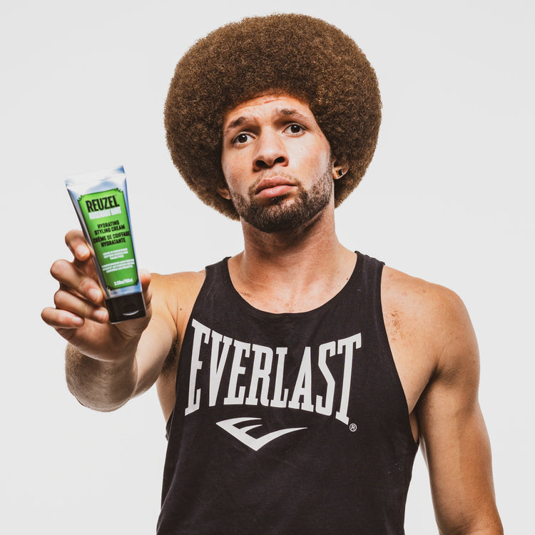 A man with gray hair and a graying beard holds up a small green dropper bottle of REUZEL rosemary mint hair and scalp serum