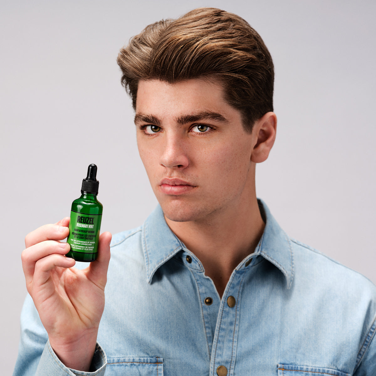 A young man with styled brown hair wearing a light blue denim shirt holds up a small green glass dropper bottle of REUZEL rosemary mint hair & scalp serum against a plain light gray studio background.