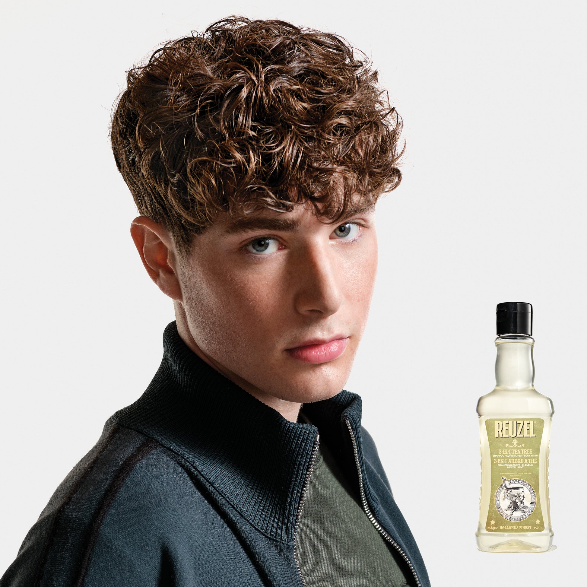 Person with styled hair next to a bottle of REUZEL 3 in 1 Shampoo on a white background