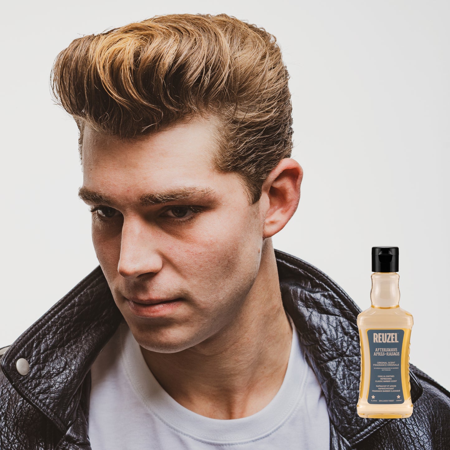 Man with styled hair wearing a leather jacket, with a bottle of Reuzel Aftershave