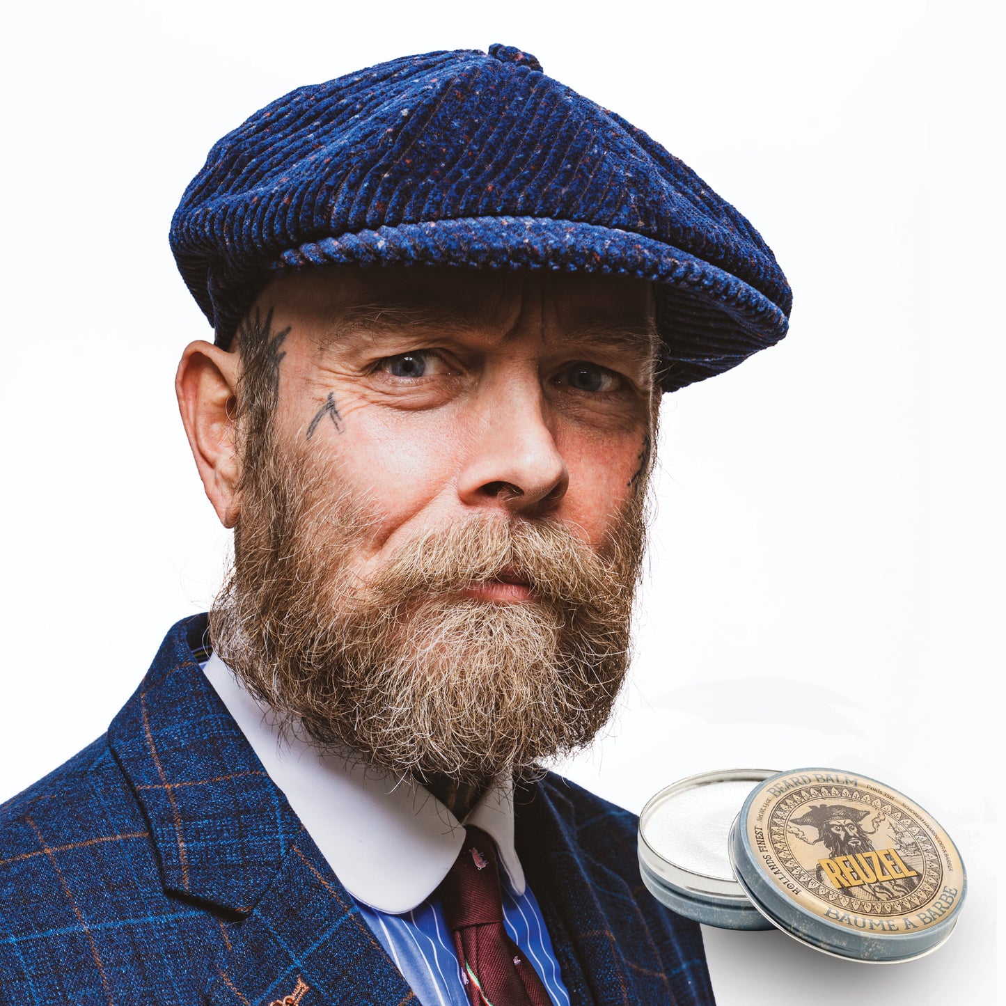 Man wearing a blue checkered cap and suit with a beard, next to a Reuzel Beard Balm on a white background