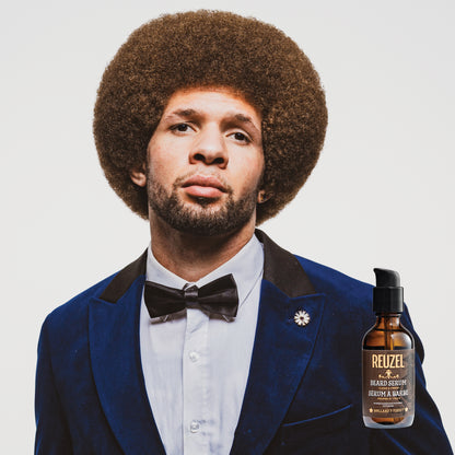 Man in a blue suit with a bow tie and a bottle of Reuzel Beard Oil