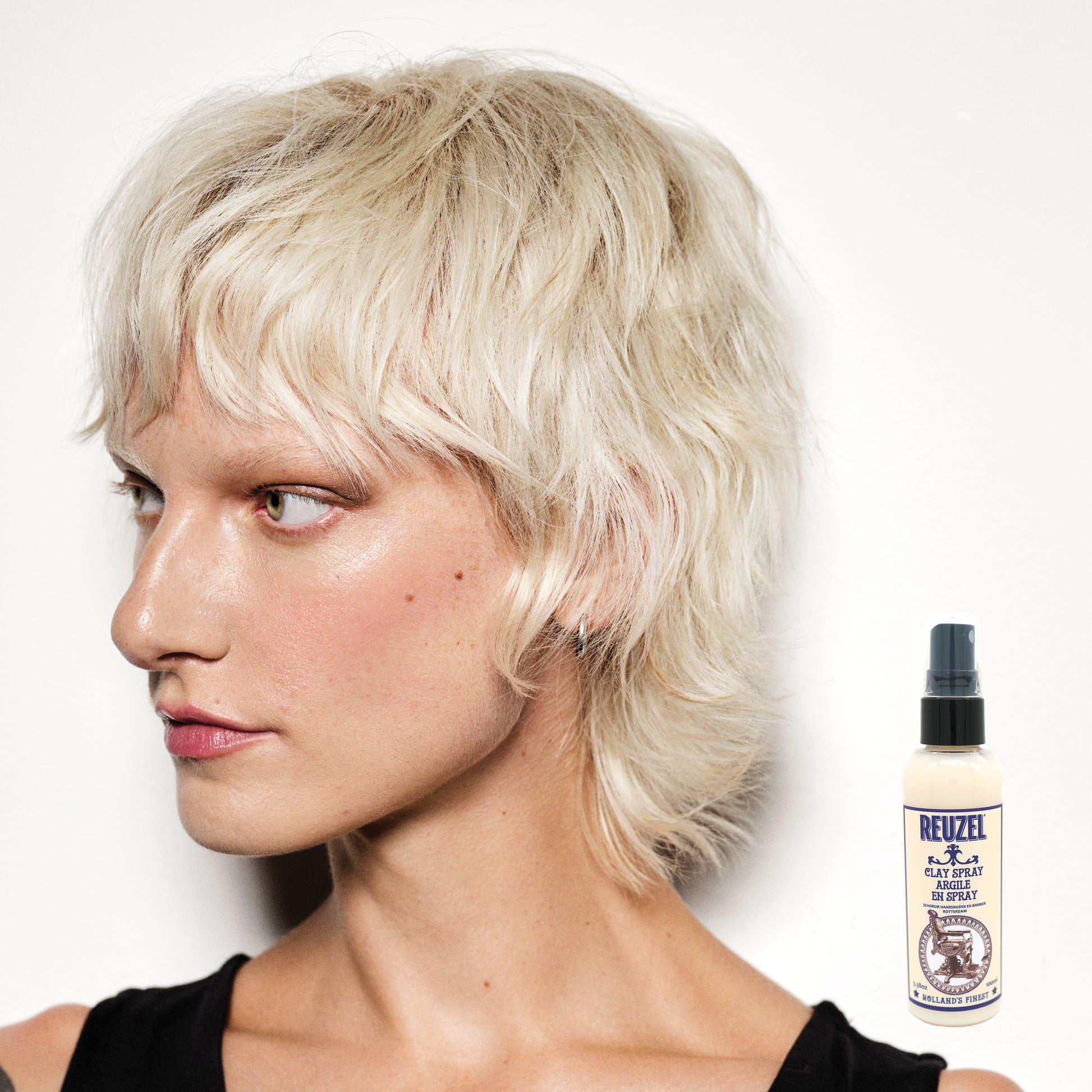 Woman with styled hair next to a bottle of Reuzel Clay Spray on a white background