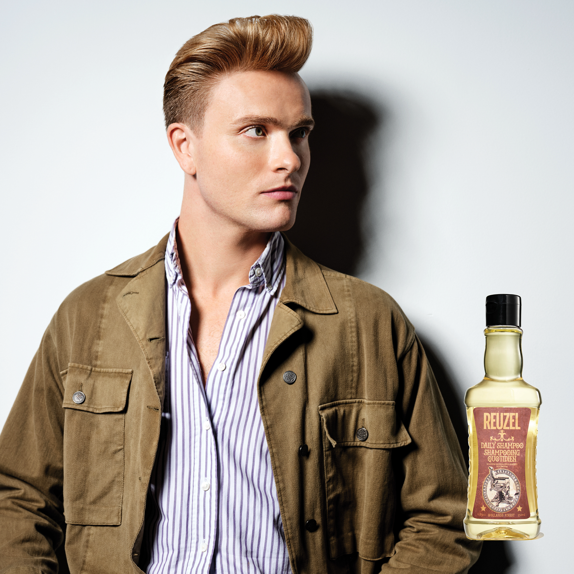 Man with styled hair and a bottle of Reuzel Daily Shampoo on a gray background