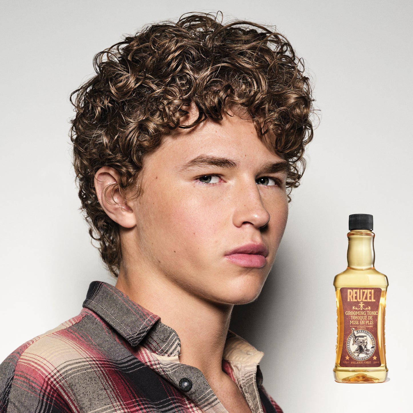 Person with styled hair next to a bottle of Reuzel Grooming Tonic on a plain background