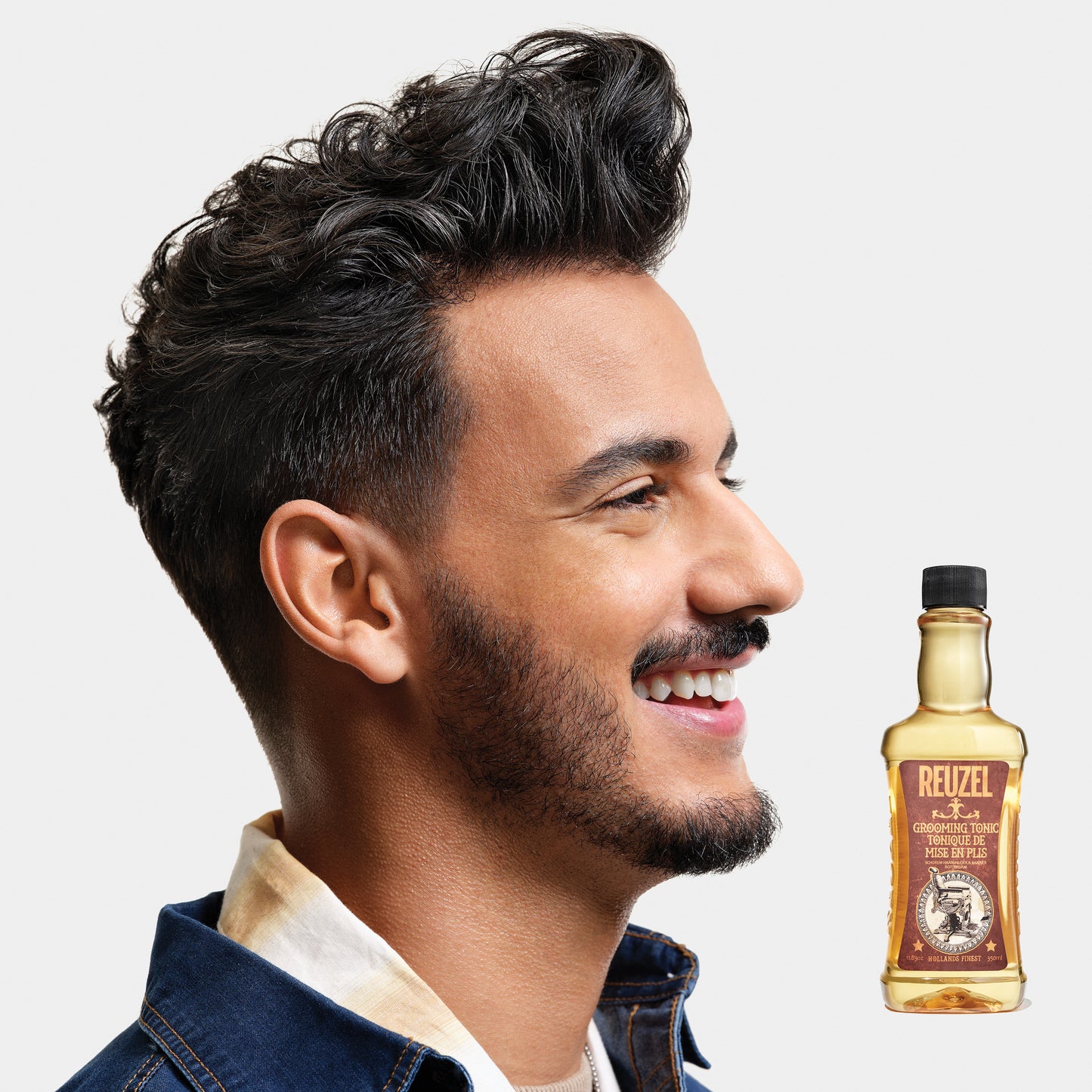 Man with styled hair next to a bottle of Reuzel Grooming Tonic on a light gray background