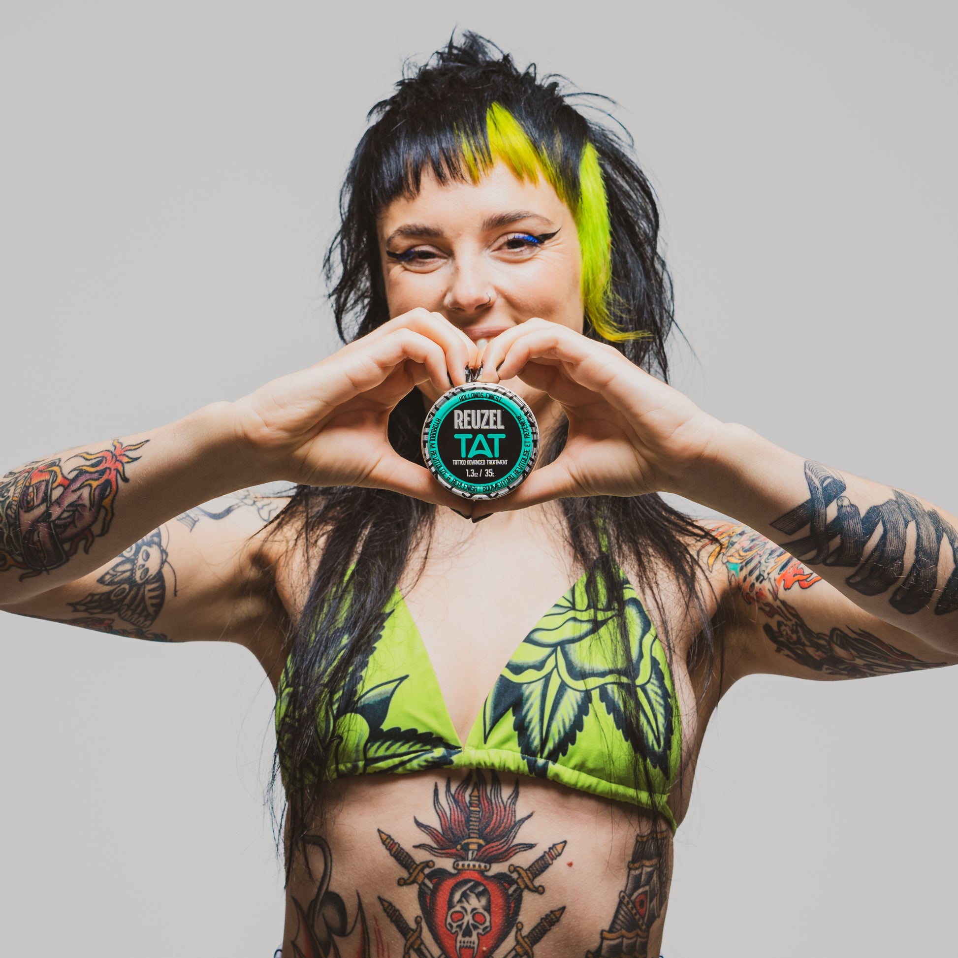 Person with tattoos holding a HYDRABALM product labeled 'Reuzel Tat' against a plain background