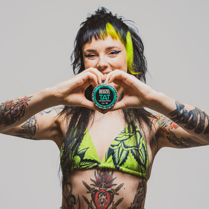Person with tattoos holding a HYDRABALM product labeled 'Reuzel Tat' against a plain background