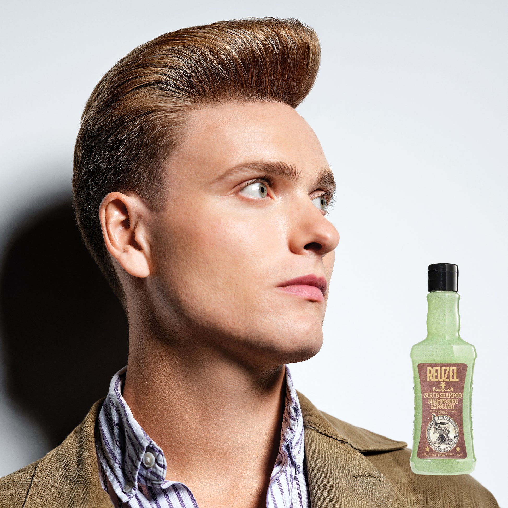 Man with styled hair next to a bottle of Reuzel Scrub Shampoo on a light gray background