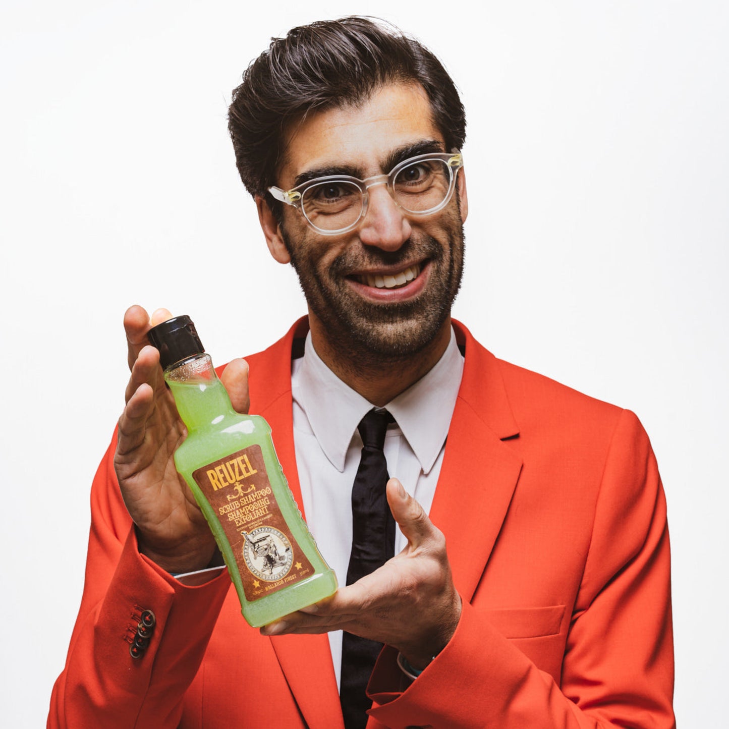 Man in a red suit holding a bottle of 'Reuzel Scrub Shampoo' against a white background