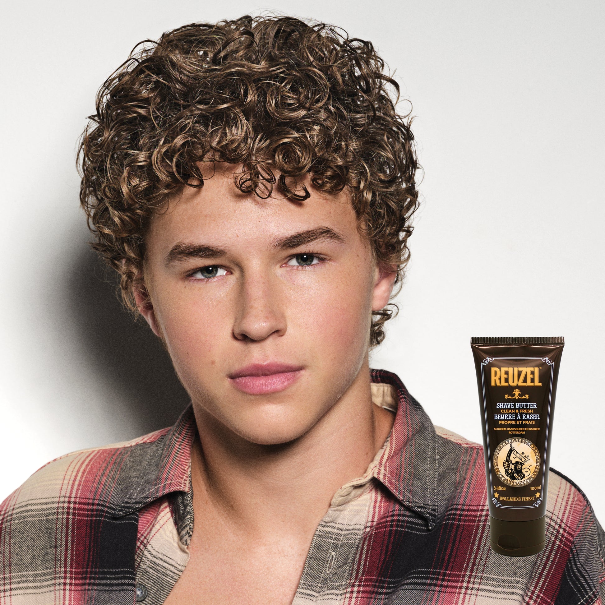 Man with curly hair wearing a plaid shirt, with Reuzel Shave Butter displayed.