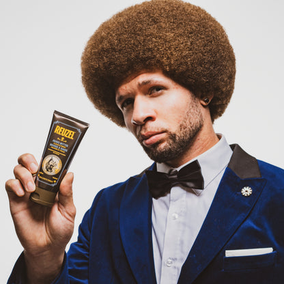 Man in a blue suit holding a tube of Reuzel Shave Butter against a white background