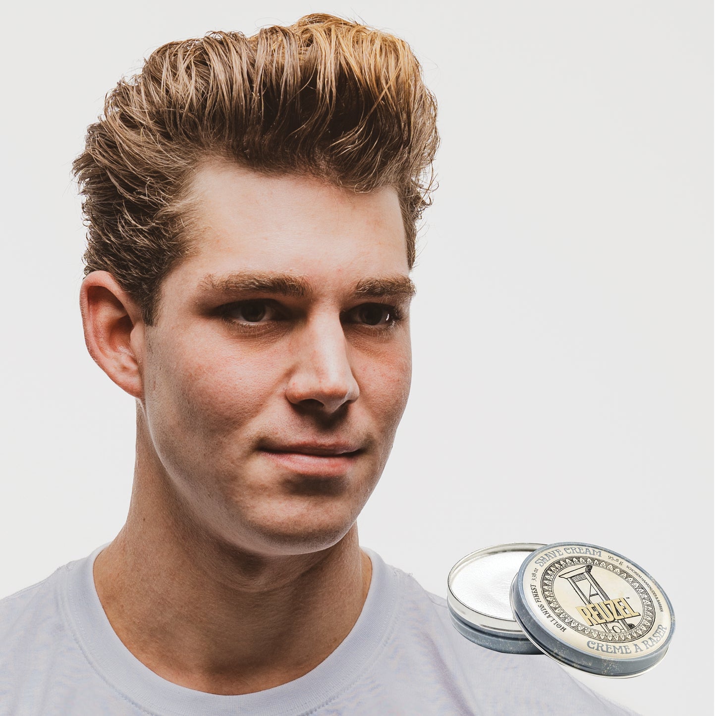 Man with styled hair next to a container of Reuzel Shave Cream on a white background