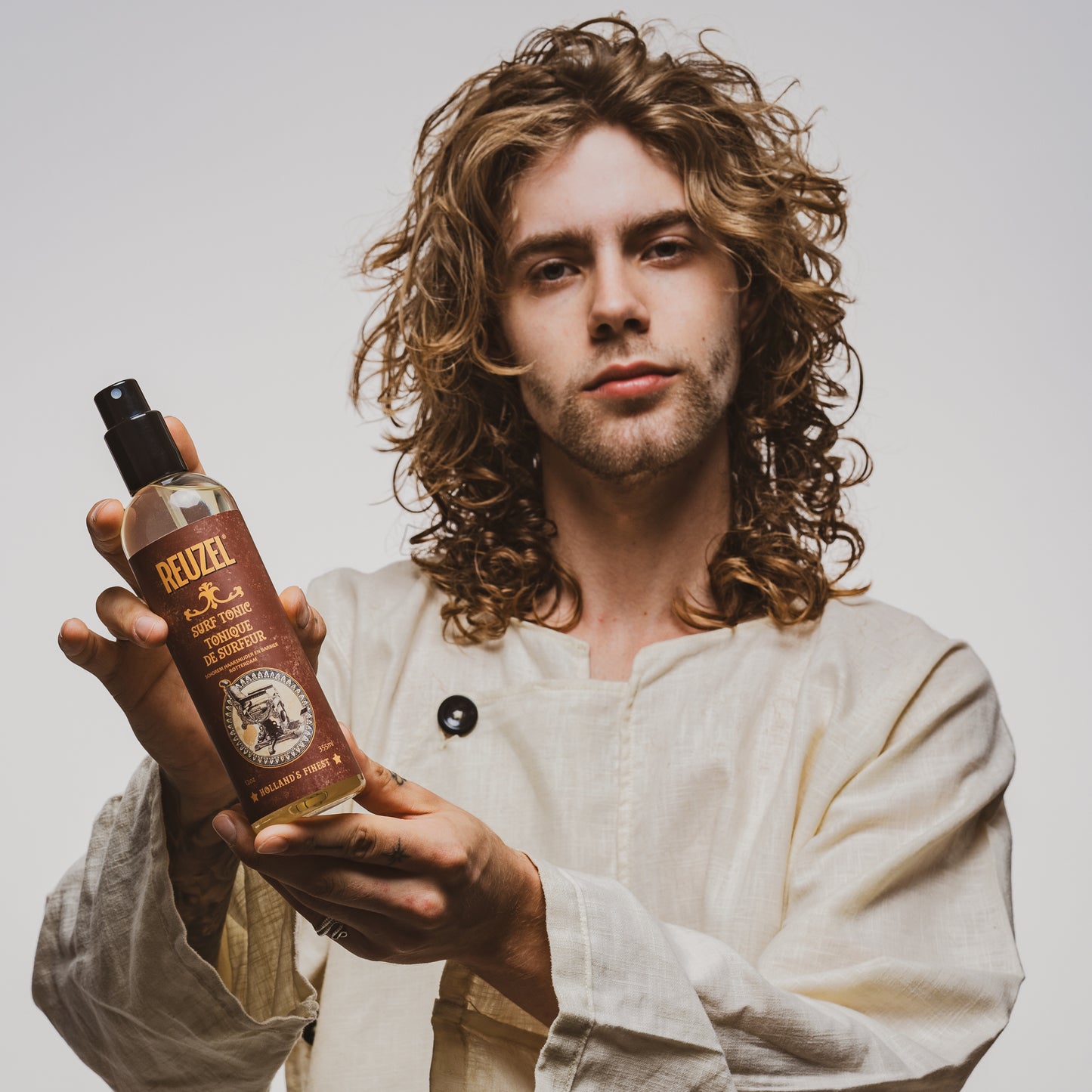 Man holding a bottle of Reuzel Surf Tonic product against a plain background