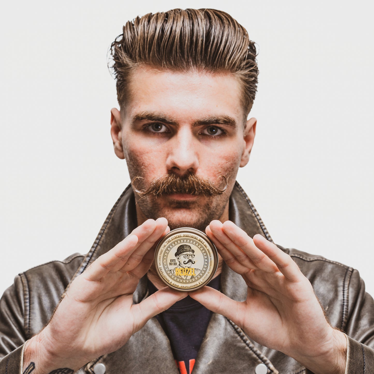 Man holding tin of Reuzel Mustache Wax, wearing a brown leather jacket against a plain background
