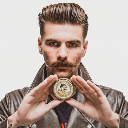 Man holding tin of Reuzel Mustache Wax, wearing a brown leather jacket against a plain background