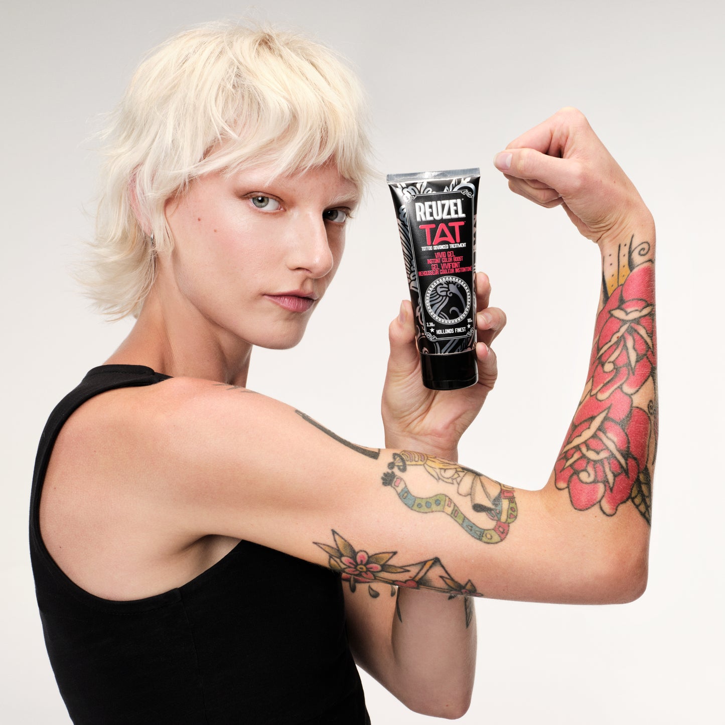 Woman with tattoos holding a tube of Reuzel TAT VIVID product against a plain background